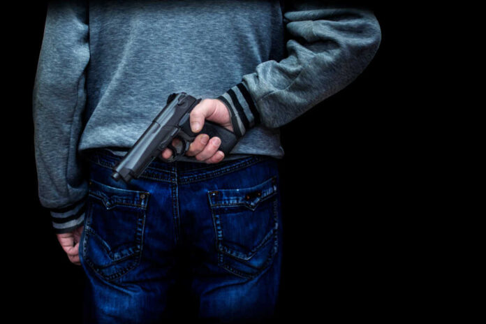 Man,Holding,A,Gun,Behind,His,Back,Against,A,Black