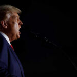 Profile view of a man passionately speaking into a microphone against a dark background