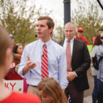 A man in a light blue shirt and red tie speaking at a rally surrounded by supporters and media