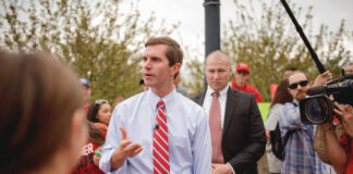 A man in a light blue shirt and red tie speaking at a rally surrounded by supporters and media