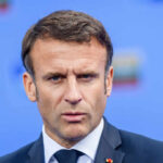 Close-up portrait of a serious-looking male political leader in formal attire against a blue background