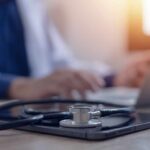 A stethoscope resting on a tablet with a blurred medical professional in the background typing on a laptop
