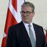 A man in formal attire standing at a podium with flags in the background