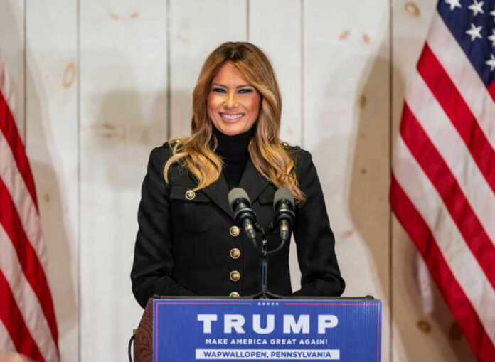 A woman in a black jacket smiling at a podium with a Trump sign and American flags in the background