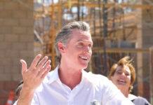 A man speaking at a construction site with a microphone, smiling, while a woman in the background laughs.