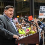 A speaker addressing a crowd at a political rally with protest signs in the background