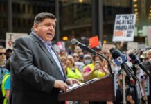 A speaker addressing a crowd at a political rally with protest signs in the background