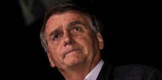 A man with a serious expression speaking into a microphone against a dark background