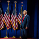 A man in a suit standing on stage in front of American flags