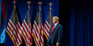 A man in a suit standing on stage in front of American flags