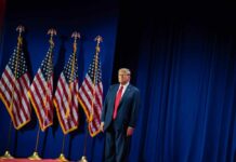 A man in a suit standing on stage next to American flags