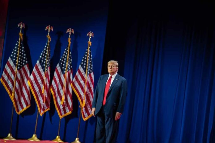 A man in a suit standing on stage next to American flags