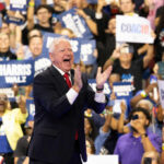A man in a suit enthusiastically claps at a political rally with a cheering crowd holding signs