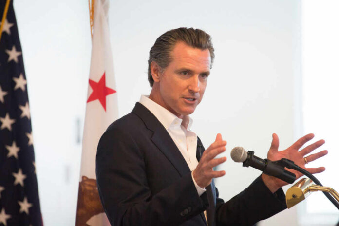 shutterstock_2503013129.jpg A man in a suit speaking into a microphone with a California flag in the background