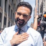 A smiling man in a light blue shirt with a tie, standing outdoors with a camera crew in the background