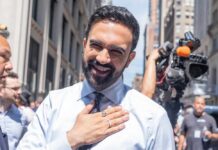 A smiling man in a light blue shirt with a tie, standing outdoors with a camera crew in the background