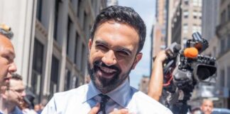 A smiling man in a light blue shirt with a tie, standing outdoors with a camera crew in the background