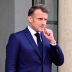 Emmanuel Macron in a suit, standing outdoors with a thoughtful expression