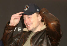 Man in a leather jacket and baseball cap smiling and adjusting his hat
