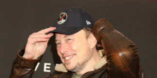 Man in a leather jacket and baseball cap smiling and adjusting his hat