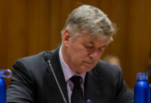 A man in a suit with a serious expression, sitting at a table with blue water bottles