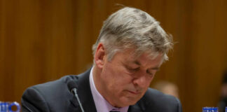 A man in a suit with a serious expression, sitting at a table with blue water bottles