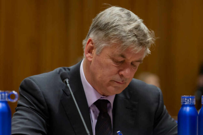 A man in a suit with a serious expression, sitting at a table with blue water bottles