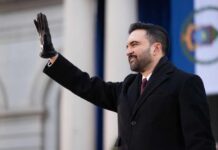 A man in a black coat and gloves waving at an outdoor event