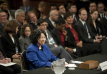 A woman in a blue suit smiling at a Senate confirmation hearing with an audience in the background