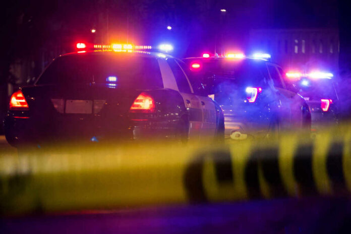 Police cars with flashing lights at a nighttime scene behind crime scene tape