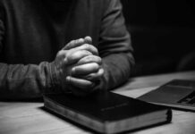 OUTRAGE: Pastor’s Bible Verse Lands Him in Court A persons hands clasped together on a desk with a bible and a laptop nearby