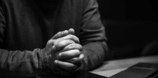 A persons hands clasped together on a desk with a bible and a laptop nearby