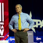 A man in a blue shirt and yellow tie stands on stage at a political conference