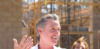 A man speaking at a construction site with a microphone, smiling, while a woman in the background laughs.
