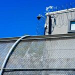 Security camera mounted above a prison fence with barbed wire against a blue sky