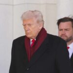 A political figure walking with a serious expression, wearing a dark coat and red scarf