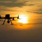 A silhouette of a drone flying against a sunset sky