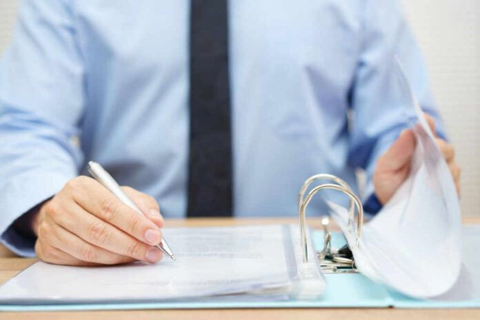 shutterstock285777377jpg A person in a blue shirt writing on a document while holding a binder