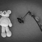 A crocheted teddy bear next to a wilted rose on a textured surface