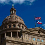 The dome of a state capitol building with flags flying against a blue sky