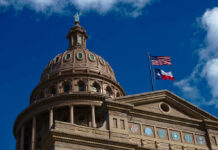The dome of a state capitol building with flags flying against a blue sky