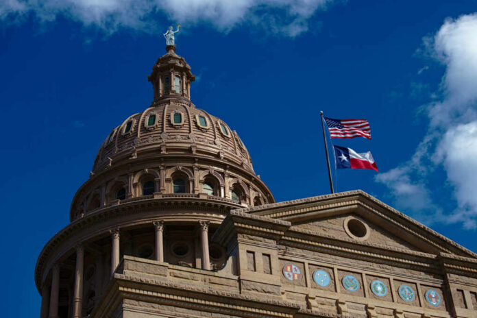 shutterstock_1673539927 The dome of a state capitol building with flags flying against a blue sky