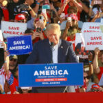 Donald Trump speaking at a political rally with supporters holding signs