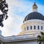 View of a state capitol dome with American and California flags against a blue sky