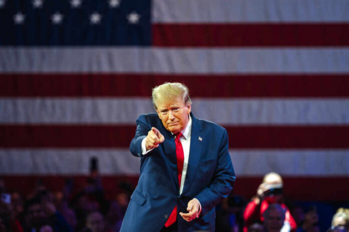 shutterstock2455396913jpg A speaker in a suit gesturing towards the audience in front of an American flag