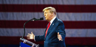A man in a suit speaking at a podium with an American flag in the background