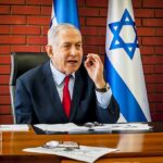 man in suit speaking at a desk with Israeli flags behind him