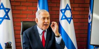 man in suit speaking at a desk with Israeli flags behind him
