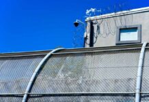 Runway Detention Hub Sparks Fury Security camera mounted above a prison fence with barbed wire against a blue sky
