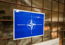 France Boosts NATO’s Northern Force NATO emblem on a blue sign attached to a rusty wire fence